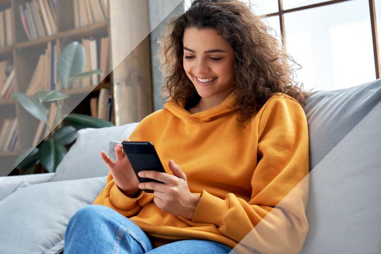 Woman in an orange sweatshirt and jeans sits on her couch and scrolls through her mobile phone